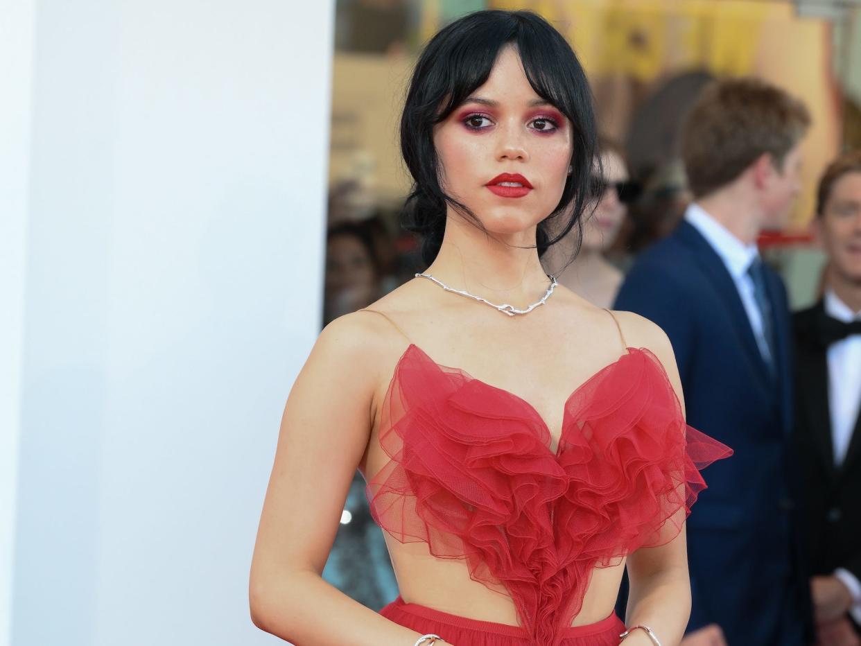 A woman with black hair wearing red eye shadow and lipstick with a silver necklace and a red dress, designed in the shape of a heart.