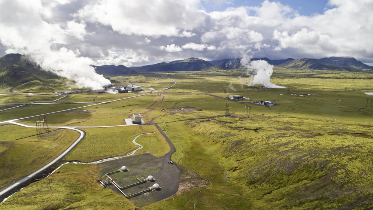 An aerial view shows a series of buildings with clouds of steam rising from them in a green landscape with mountains in the background. 