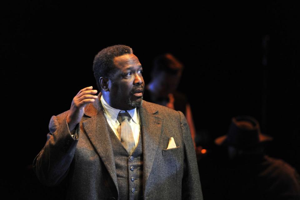 wendell pierce wearing a gray suit, standing on a darkened stage, looking off camera and holding his right hand in the air