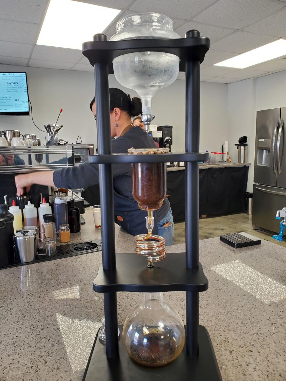 Cold water drips from the top glass globe over the ground coffee in the center glass globe into the bottom third globe of the Yama Cold Brew Tower at Go Make Coffee Co. as barista Michelle Keefaw makes coffee drinks for customers.