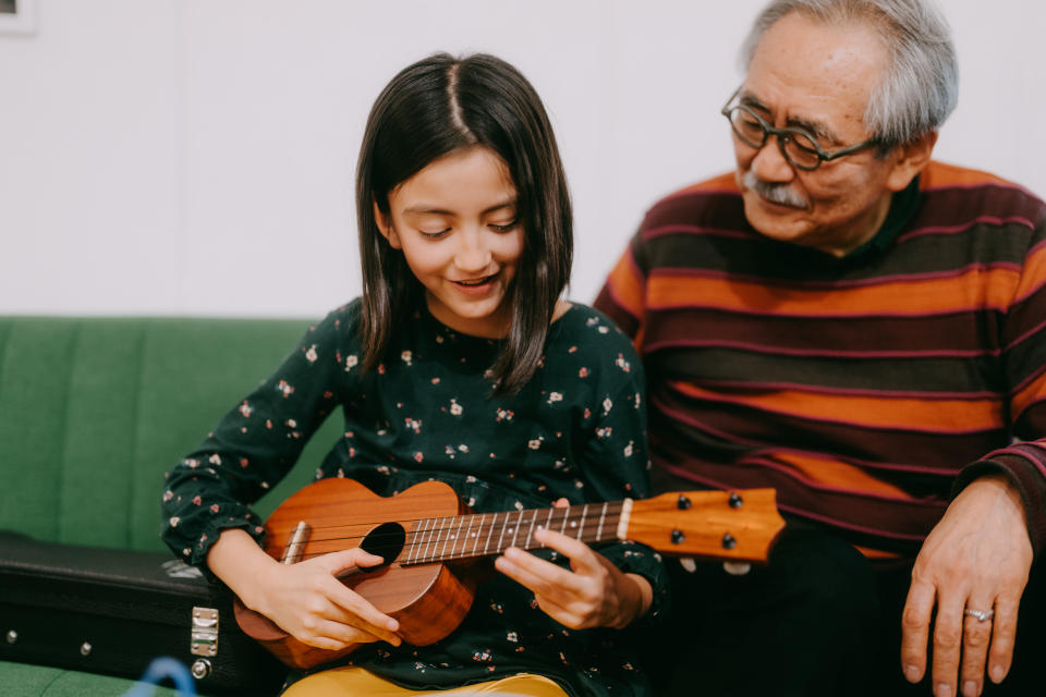 Cute young girl playing ukulele with her grandfather at home