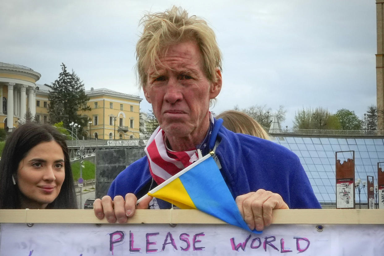 Ryan Wesley Routh, holding a small Ukrainian flag, stands near several buildings among a few other people with his hands on top a banner that reads, in part: Please world.