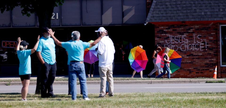 As people from New Way Church pray on the median separating Eastpointe from Detroit on 8 Mile Road, members of Gamp Camp Escorts use umbrellas to shield a client from them as she walks out of the Eastland Women's Clinic in Eastpointe on July 2, 2022. Gamp Camp Escorts have seen a rise in volunteers signing up to help since the reversal of Roe vs Wade in late June.