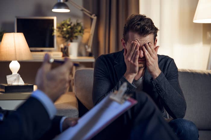 A man sits on a sofa covering his face with his hands, looking distressed. Another person holds a clipboard, suggesting a counseling or therapy session