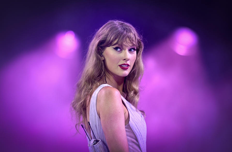 Taylor Swift, on stage, looks over her shoulder with a slight smile. She wears a sleeveless dress, with soft lighting shining on her