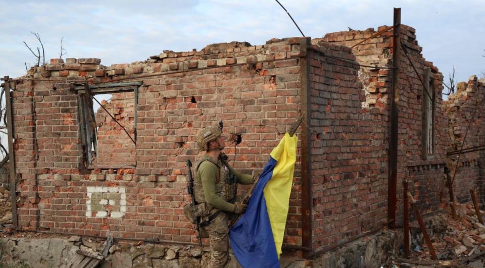 A soldier holds the Ukrainian flag in front of a destroyed building in the village of Andriivka.