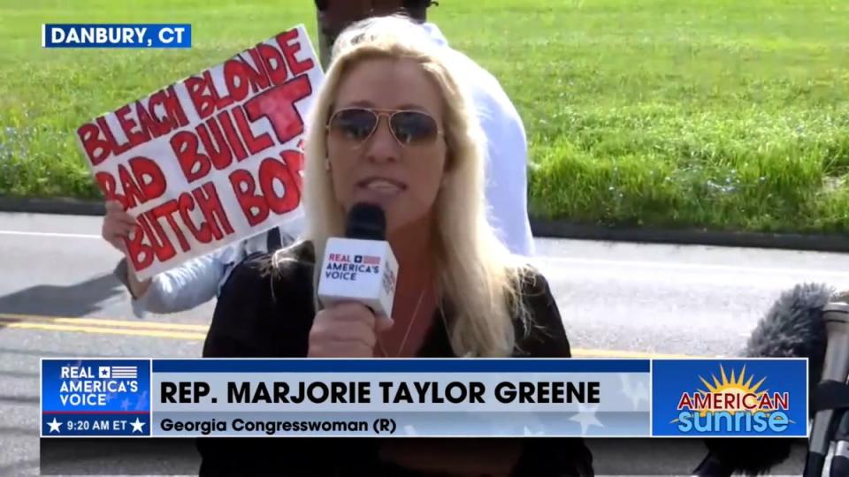 Majorie Taylor Greene speaks outside a Danbury, Connecticut, prison as Steve Bannon turns himself in. A protestor held up a sign mocking the Georgia representative during her TV appearance (Real America's Voice)