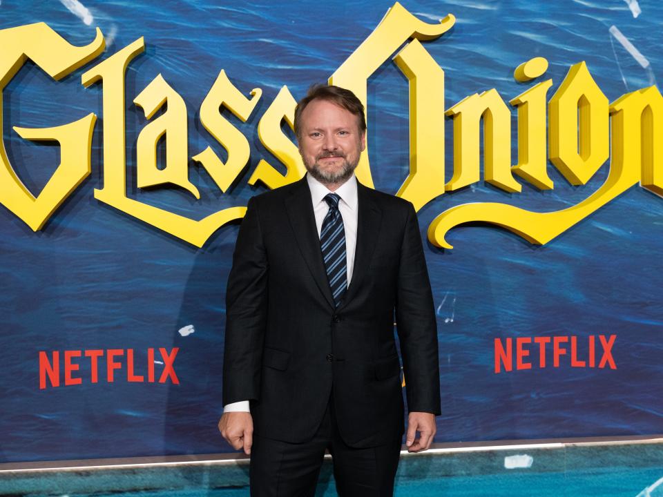 Rian Johnson in a suit and tie in front of a Glass Onion step and repeat