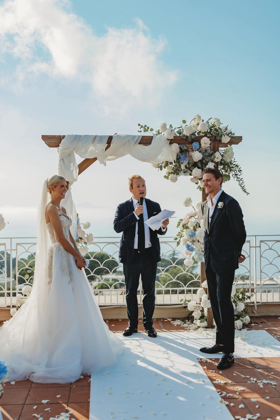 A bride and groom say their vows in front of a floral archway and the ocean.