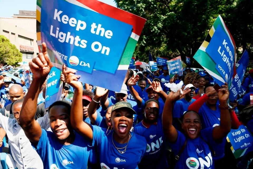 Crowd with signs reads "Keep the Lights On" and "Vote DA", showing support for a political campaign