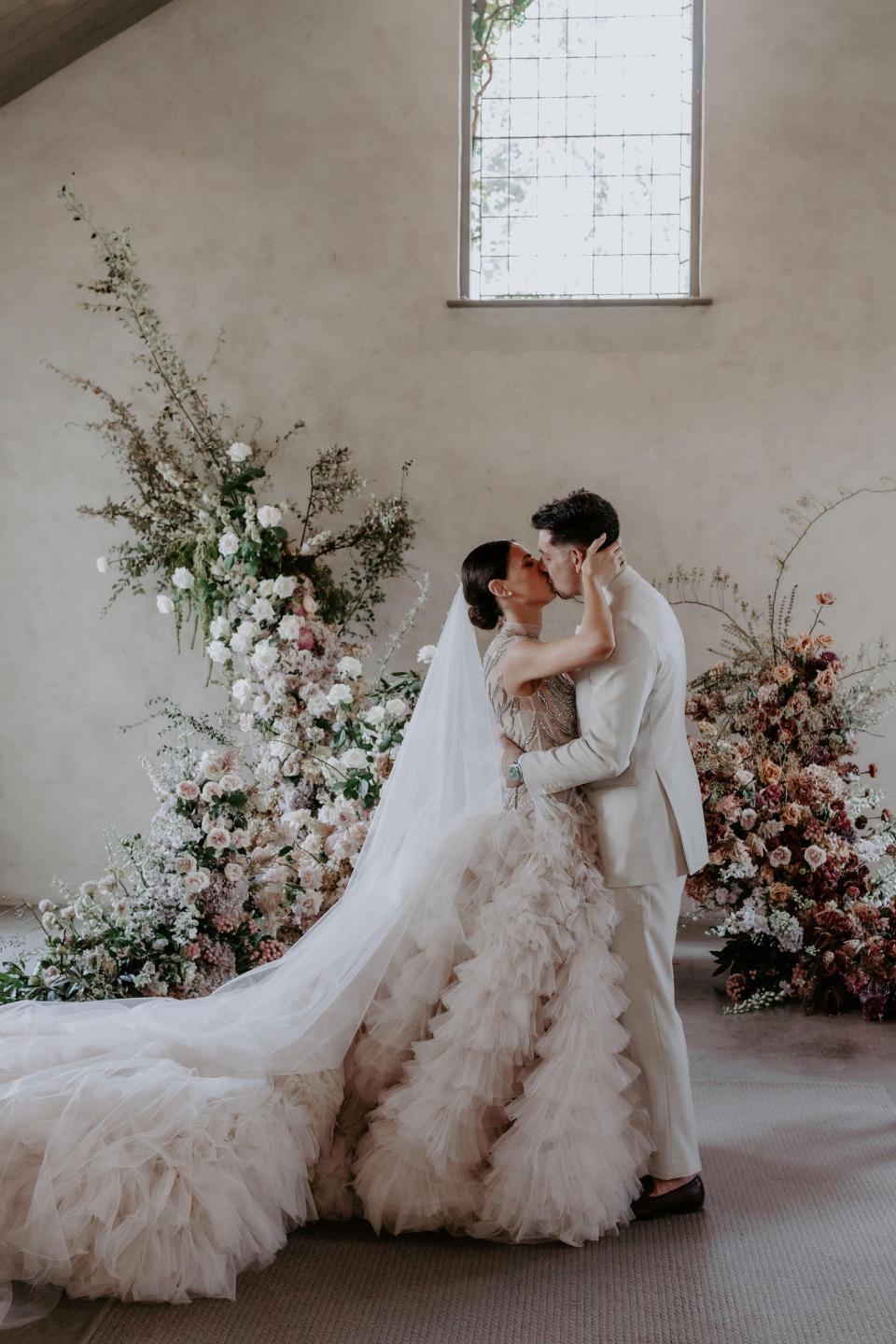 A bride and groom kiss in front of an ornate flower arrangement.