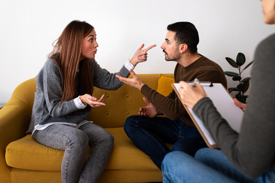 A man and a woman are arguing on a yellow couch while a person with a clipboard takes notes