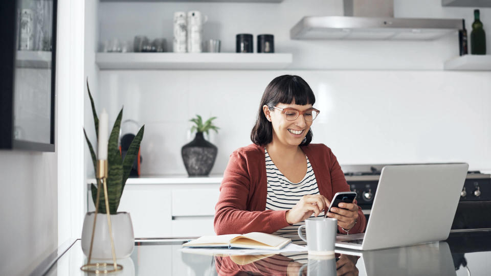 Shot of a young woman using her cellphone while sitting at home with her laptop.