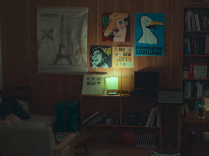 a teenage boy's bedroom with french-themed, possibly vintage posters on the back wall. the teenage boy is standing on the right, behind a desk strewn with papers