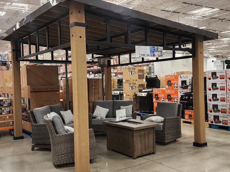 A wooden gazebo with a metal roof and detailing with a patio set of gray chairs and a brown fire pit table underneath on the floor at Costco