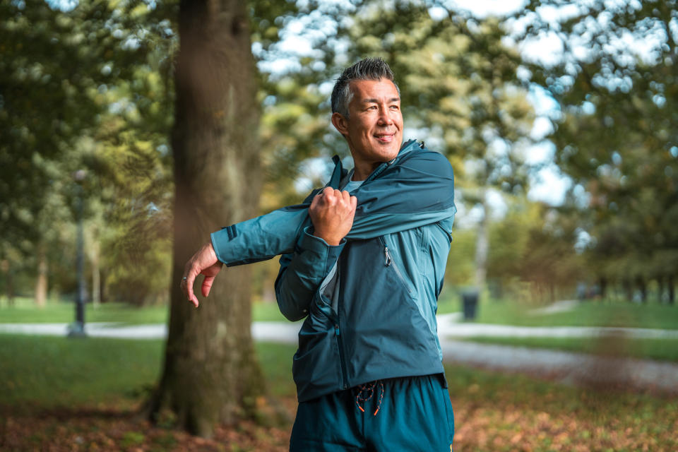 A person stretches their arm while wearing athletic clothing in a park surrounded by trees