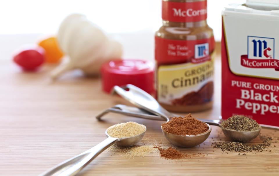 Two jars of McCormick brand cinnamon and black pepper on a wood table, with spoons full of various spices and some raw herbs.