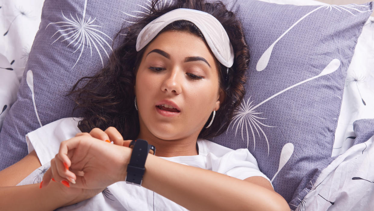  Young woman with sleep mask checking sports watch in bed. 