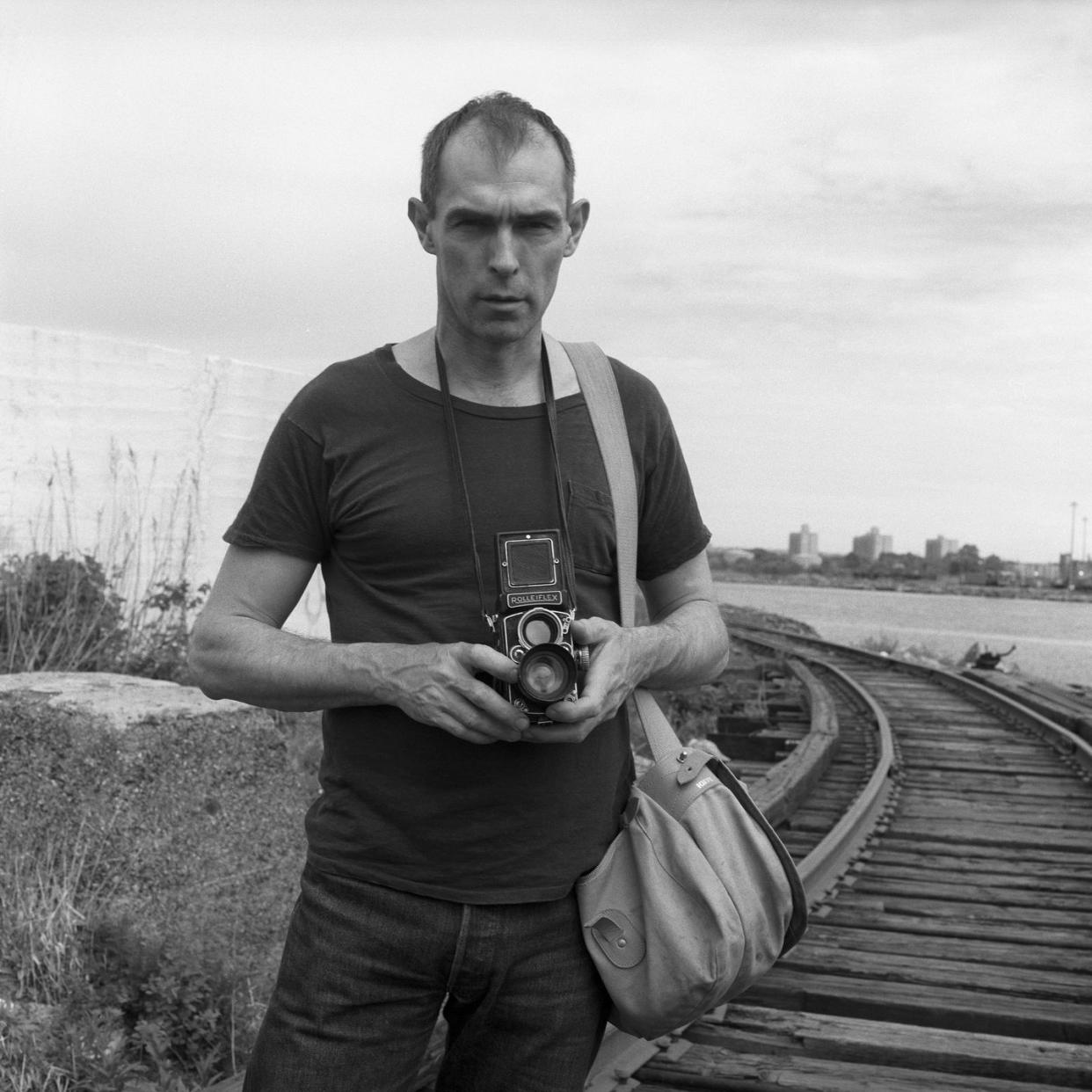 peter hujar stands holding a camera in front of a rail track in a black and white image