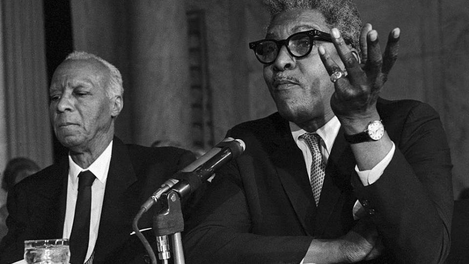 bayard rustin gesturing with his left hand a he speaks to a government committee behind a microphone