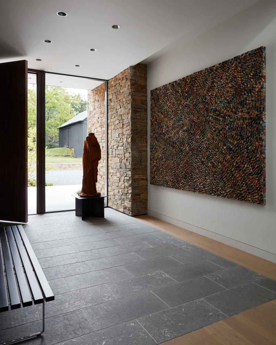 Natural materials—a walnut front door, a fieldstone wall, a stone foyer—provide a smooth transition from the outdoors. A piece by Don Morris adds more texture with its use of folded comic books. A monk figure in a saffron robe stands guard.