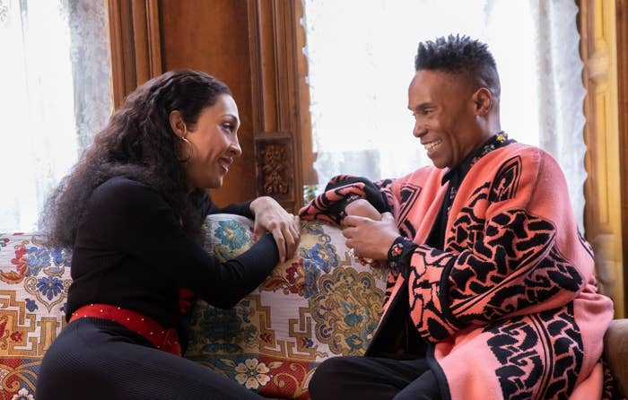 Billy Porter in a patterned coat and Michaela Jaé Rodriguez in a black outfit with red accents sit on a couch, smiling and holding hands