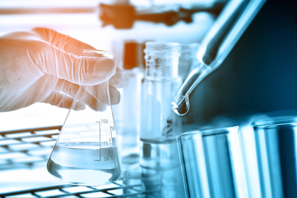 Hand holding beaker with test tubes, droppers, and other glassware in the foreground and background