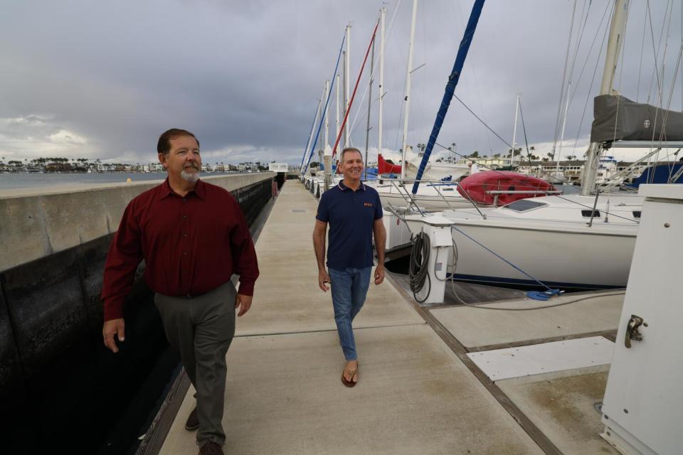 A man in a red long-sleeved shirt and gray pants, left, and another in dark blue polo shirt and jeans, walk on a dock