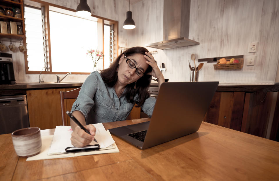 Woman sits at kitchen table in front of her laptop, looking stressed. (Source: Getty)