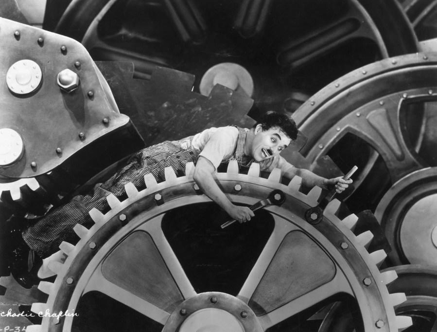 British actor and director Charlie Chaplin wearing overalls and holding a wrench, sits on an enormous set of gears in a still from Chaplin's film, 'Modern Times'.  (Photo by Hulton Archive/Getty Images)