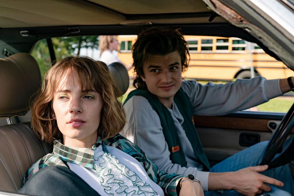 maya hawke and joe keery as robin and steve in stranger things, sitting in a car with steve at the wheel. they're both looking out the passenger side window at something