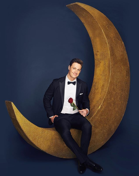 A photo of The Bachelor Australia Matt Agnew wearing a tuxedo, holding a red rose and sitting on a crescent moon.