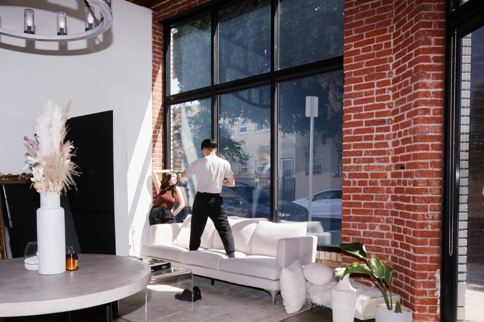 Photographer David Suh stands on a couch while photographing a client leaning against a window.