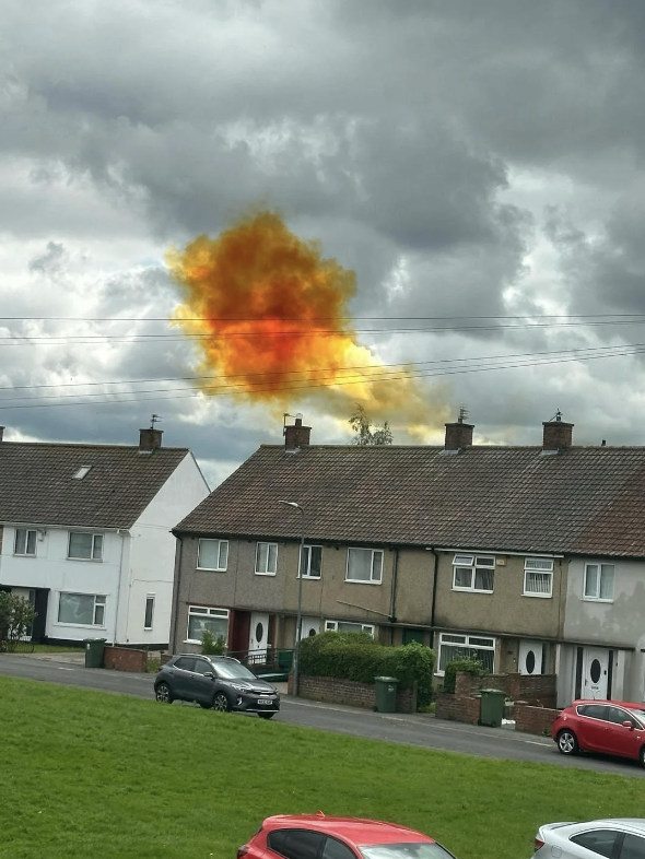 Bright orange and yellow smoke plume rises above a row of residential houses, with dark clouds in the sky