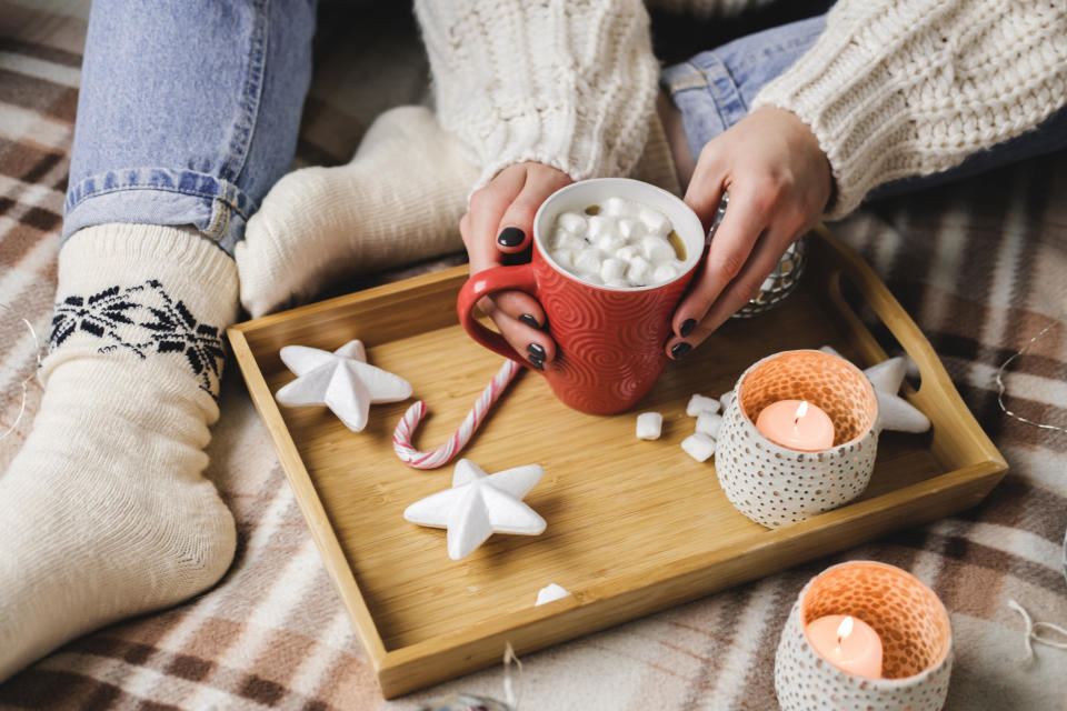 Young woman sits on plaid in cozy knitted woolen sweater and holds cup of cocoa with marshmallows. Hygge New Year, cozy Christmas, preparation for holidays. Wooden tray witch candles, stars, balls