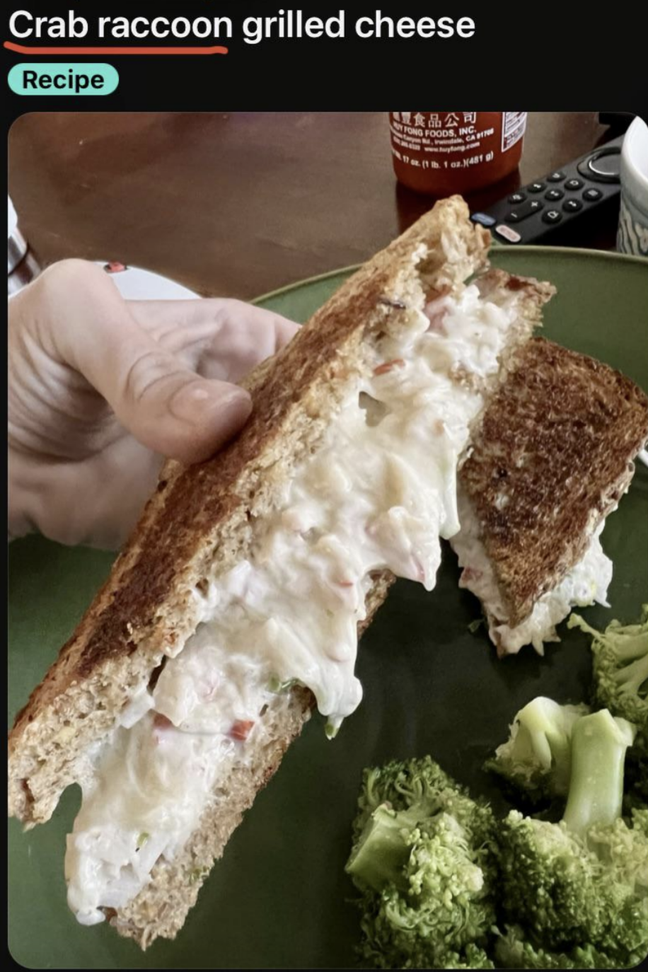 Hand holding a grilled cheese sandwich filled with crab meat mixture. A green plate with broccoli is also visible. A hot sauce bottle in the background