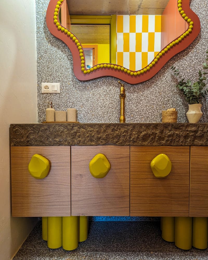 Bathroom with wavy orange and yellow mirror, wood cabinets with stone slab counter and yellow irregular shaped knobs