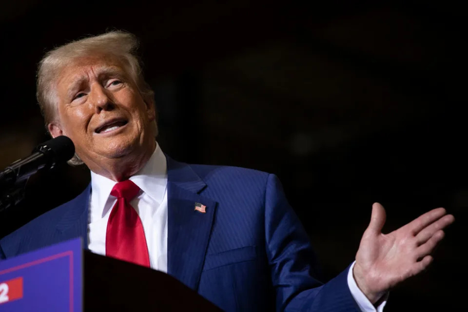 Donald Trump speaking at an event, wearing a suit and red tie, gesturing with his right hand near a microphone