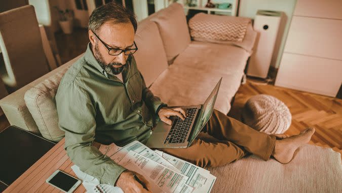 Mature man sitting on sofa in living room with laptop and financial reports, doing his monthly budget.