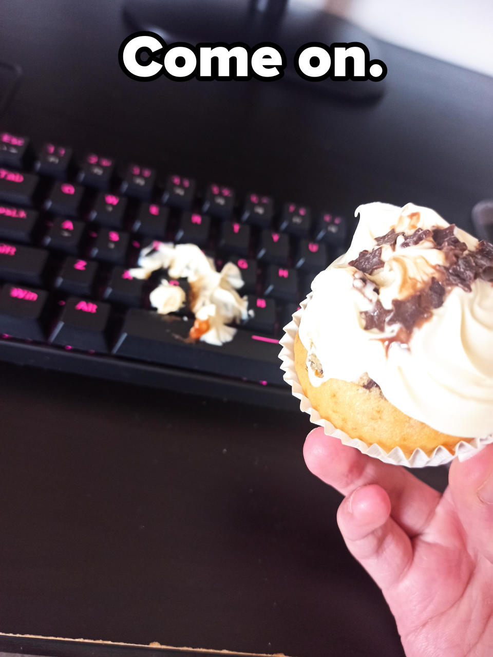 A hand holding a cupcake with frosting, in front of a keyboard with some spilled frosting on it