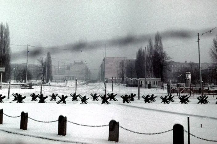 Anti-tank obstacles line a snowy field in front of large buildings in the distance.