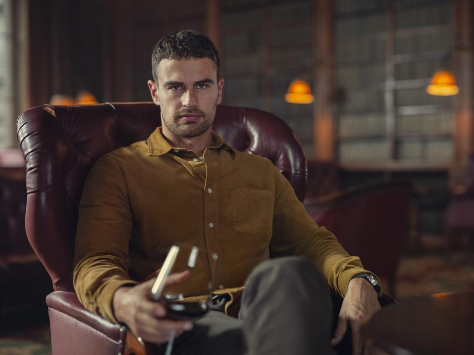 theo james as eddie horniman, sitting in a red leather chair and holding a glass of red wine