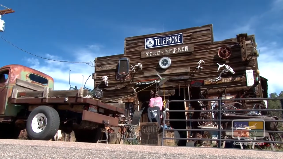 Old truck parked in front of a rustic building with eclectic decor and signs stating "Telephone" and "Tire Repair."