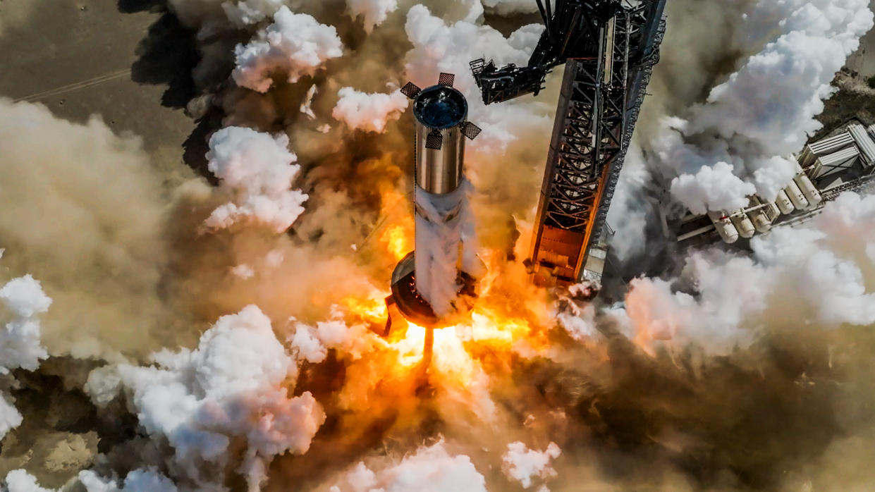  Flames billow from the base of a large silver rocket that's sitting on a test stand. 