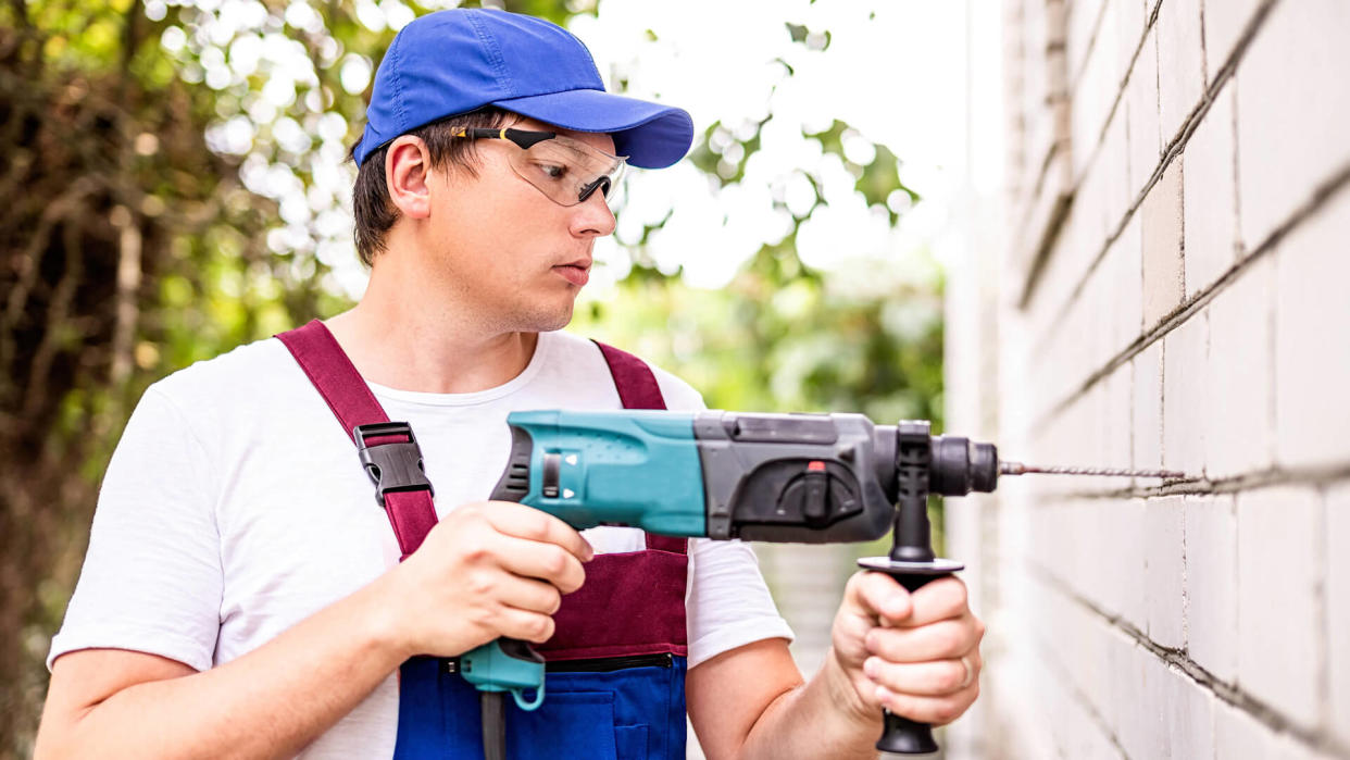 Building worker in in protection glasses and uniform with perforator drilling the wall outdoors.