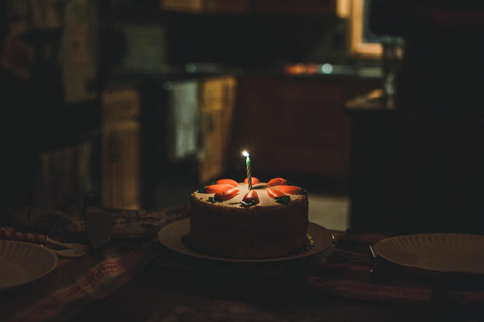 A round carrot cake sits alone on a table between two plates in a dark room with one clit birthday candle sitting in the middle of it.
