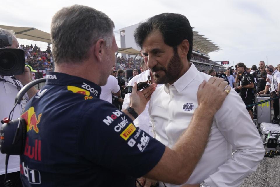 A man wearing race team clothing speaks to a man wearing a white buttondown with a logo that reads "FIA."