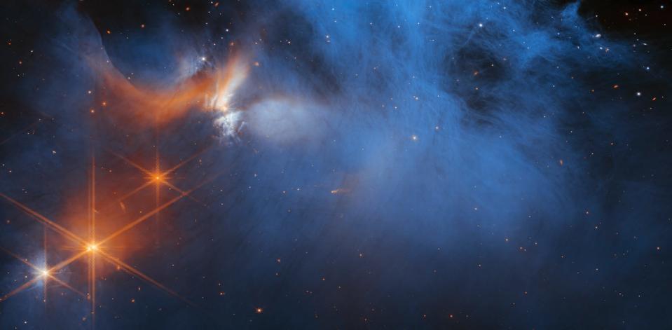 This image by NASA’s James Webb Space Telescope’s Near-Infrared Camera (NIRCam) features the central region of the Chamaeleon I dark molecular cloud, which resides 630 light years away. The cold, wispy cloud material (blue, center) is illuminated in the infrared by the glow of the young, outflowing protostar Ced 110 IRS 4 (orange, upper left). The light from numerous background stars, seen as orange dots behind the cloud, can be used to detect ices in the cloud, which absorb the starlight passing through them.An international team of astronomers has reported the discovery of diverse ices in the darkest regions of a cold molecular cloud measured to date by studying this region. This result allows astronomers to examine the simple icy molecules that will be incorporated into future exoplanets, while opening a new window on the origin of more complex molecules that are the first step in the creation of the building blocks of life.