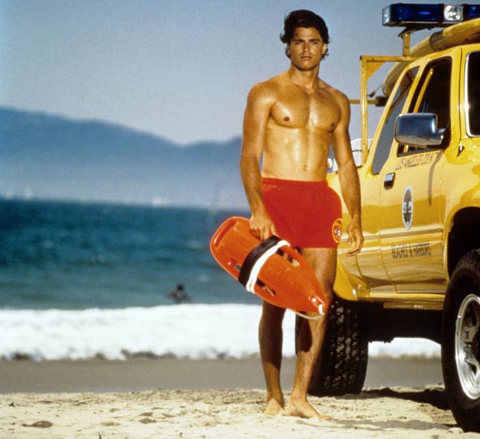 A shirtless male lifeguard standing on a beach holding a rescue float, next to a lifeguard vehicle with the ocean in the background