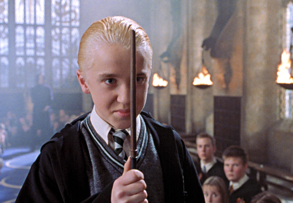 Tom Felton as Draco Malfoy, wearing a school robe and holding a wand in a scene from the "Harry Potter" series, with slicked back, platinum hair